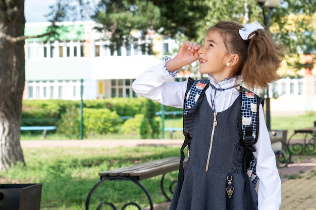 Garota alegre e engraçada em uniforme escolar com laços brancos no pátio da escola aponta com o dedo De volta à escola, 1º de setembro Aluno feliz com uma mochila Classe primária do ensino fundamental