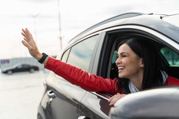 Garota alegre está acenando para alguém enquanto está sentado no carro novo com prazer. Ela está sorrindo com prazer e olhando para longe. Conceito de passageiro