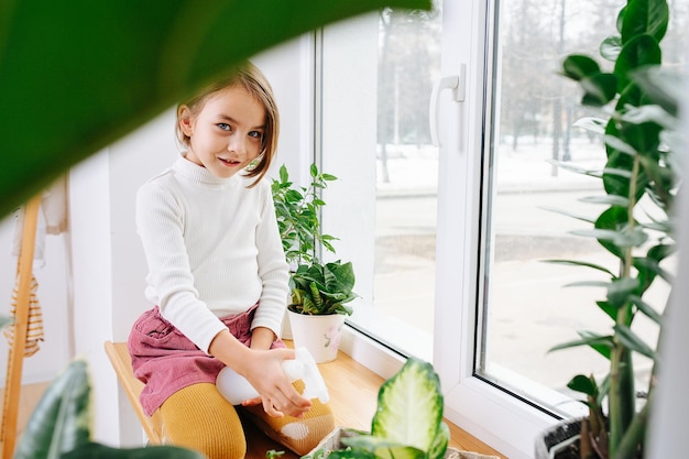 Garota alegre sentada no peitoril da janela pulverizando plantas da casa com água