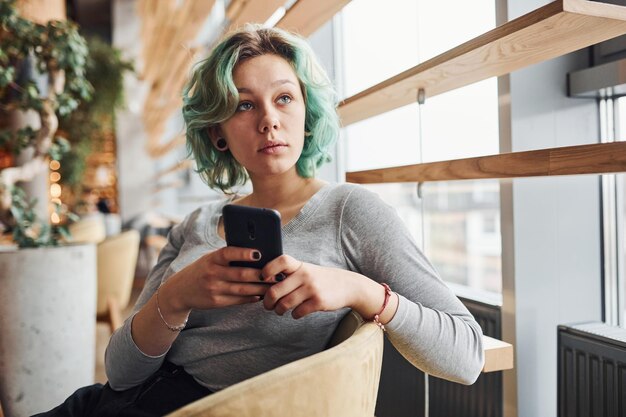 Garota alternativa com cabelo verde sentado dentro de casa durante o dia com telefone nas mãos.