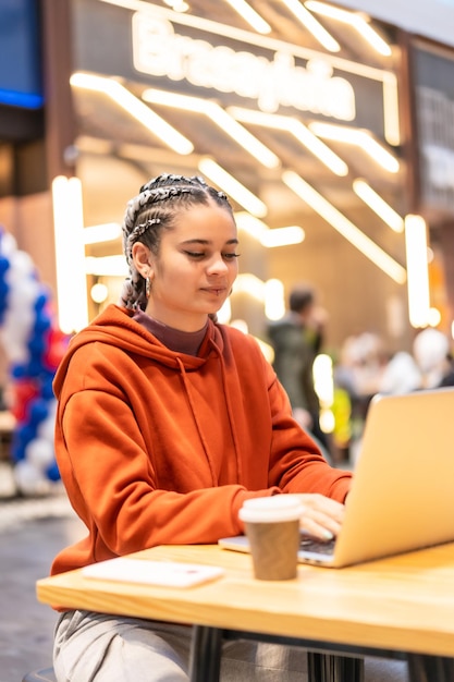 Foto garota alternativa com tranças brancas com um computador em um shopping center, enviando um e-mail com um café quente nas mãos