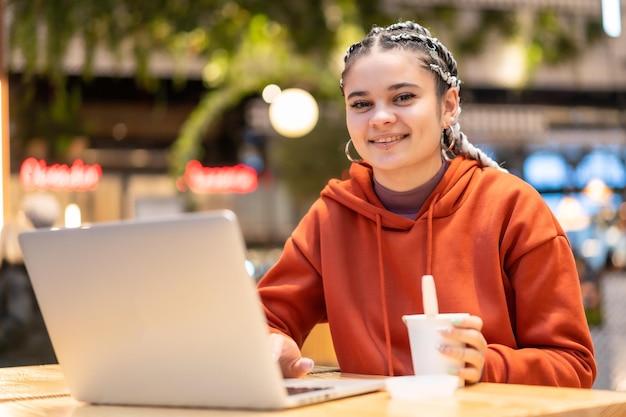 Garota alternativa com tranças brancas com um computador em um shopping center, fazendo uma videochamada com um café quente nas mãos