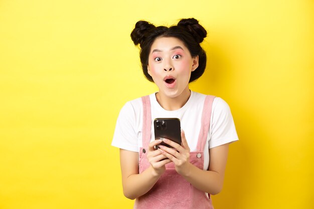 Garota animada com maquiagem glamourosa e roupas de verão, arfando e espantada, verificando a oferta de internet no celular, parada na amarela