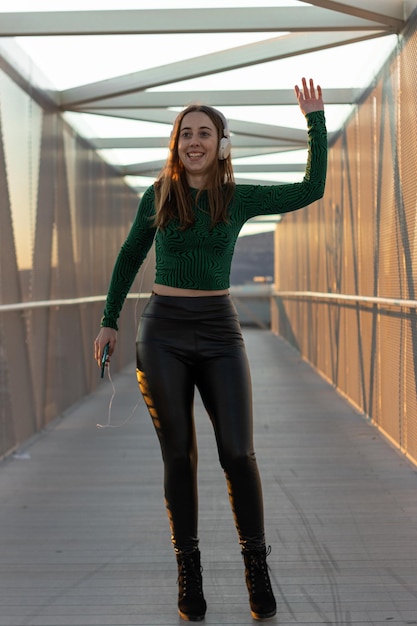 Garota animada dançando e ouvindo música com fones de ouvido e telefone inteligente e gesto de onda dançando na rua no pôr do sol Retrato vertical