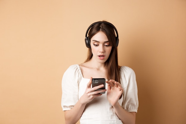 Garota animada ouvindo música em fones de ouvido e olhando para smartphone maravilhada lendo notícias incríveis encontradas ...