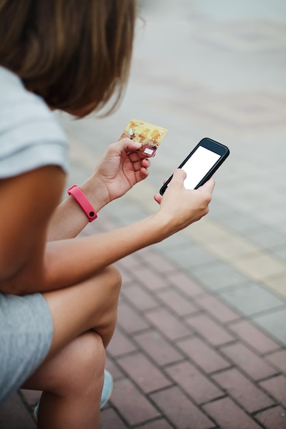 Garota anônima navegando no smartphone enquanto segura o cartão e está sentado do lado de fora fazendo compras online