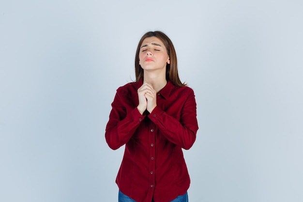 Foto garota apertando as mãos em gesto de oração em camisa cor de vinho e olhando esperançosa.