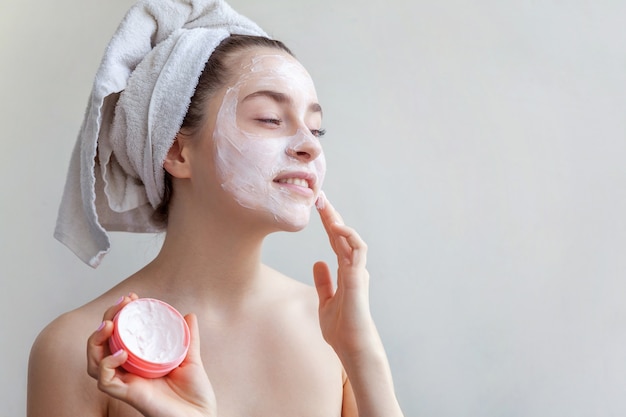 Garota aplicando máscara facial branca isolada no fundo branco. Jovem de toalha na cabeça com máscara nutritiva branca ou creme no rosto. Conceito de beleza natural e cosmetologia do spa de tratamento de cuidados da pele.