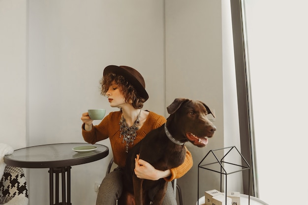 Garota atraente abraça um lindo cachorro marrom e bebe café em um café bem iluminado