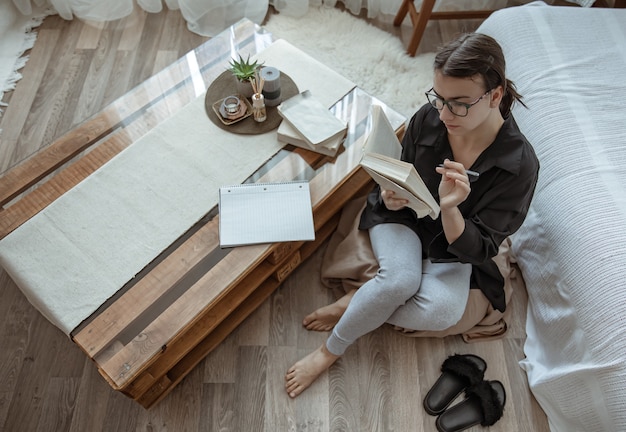 Garota atraente com óculos lê um livro sentado em um pufe em casa.