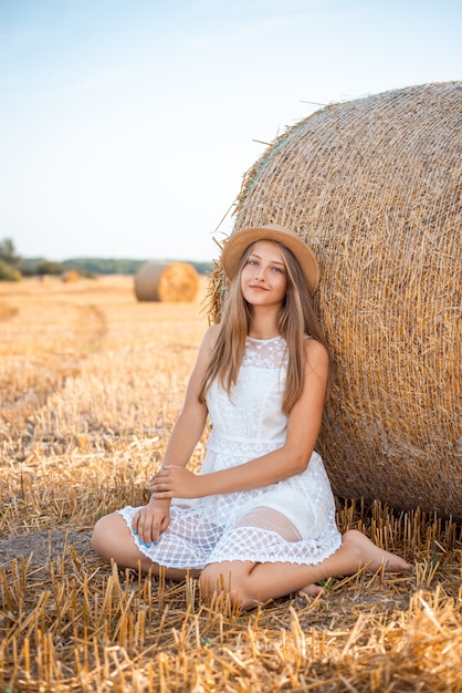 Garota atraente com os pés descalços em um vestido branco e um chapéu de palha perto de um fardo de palha no campo