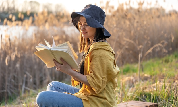 Garota atraente com um chapéu lendo um livro sobre a natureza ao pôr do sol