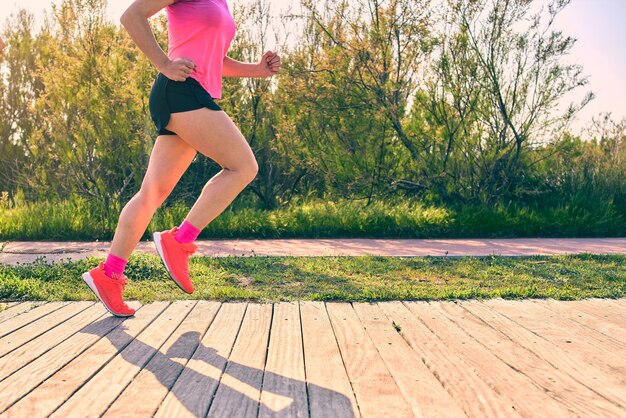 Garota atraente correndo. Aproxime-se dos pés e pernas durante o treinamento da técnica de corrida para evitar lesões. Camisa rosa e tênis. Caminho de madeira.