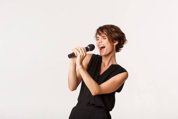 Garota atraente despreocupada cantando no microfone, se apresentando, se divertir no karaokê, em pé sobre o branco.