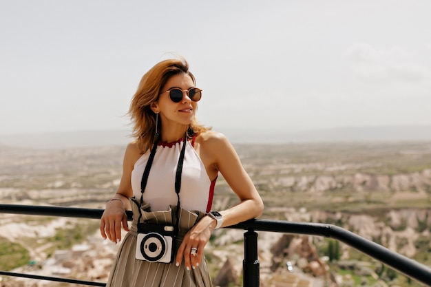 Garota atraente e bonita com cabelo voador, usando óculos escuros e segurando uma câmera retro, olhando para longe e sorrindo na paisagem montanhosa