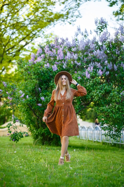 Garota atraente elegante e sorridente com cabelo comprido caminha no parque, usando um vestido marrom quente com um chapéu. Mulher jovem de aparência europeia