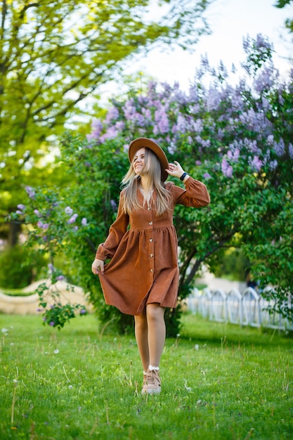 Garota atraente elegante e sorridente com cabelo comprido caminha no parque, usando um vestido marrom quente com um chapéu. Mulher jovem de aparência europeia