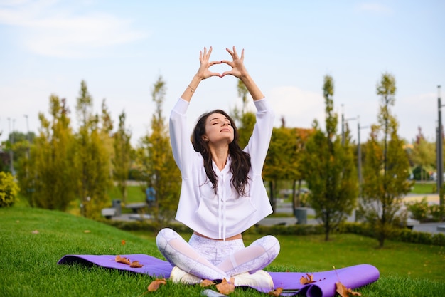 Garota atraente em roupas esportivas brancas fazendo ioga mudra de dedos com as mãos levantadas, sentada no tapete no parque da cidade, exercício ao ar livre