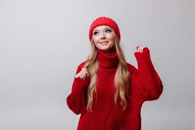 Garota atraente em um suéter vermelho de inverno e chapéu em um fundo cinza com maquiagem brilhante