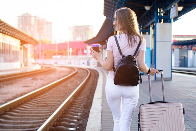 Garota atraente está de pé com bagagem na estação e esperando o trem, o aluno está indo em uma viagem, ela está caminhando ao longo da plataforma com café, cópia espaço