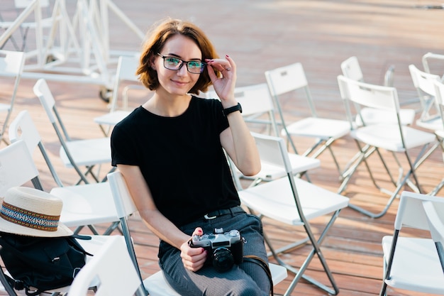 Garota atraente hipster de óculos segurando uma câmera retro nas mãos enquanto está sentada em uma cadeira ao ar livre