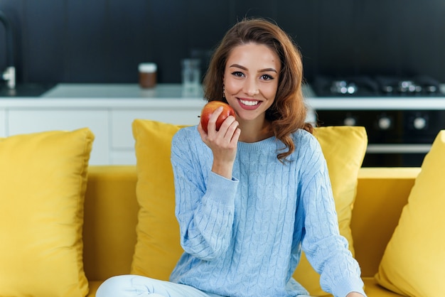 Garota atraente mantém a maçã vermelha fresca enquanto está sentado no sofá amarelo na cozinha elegante.