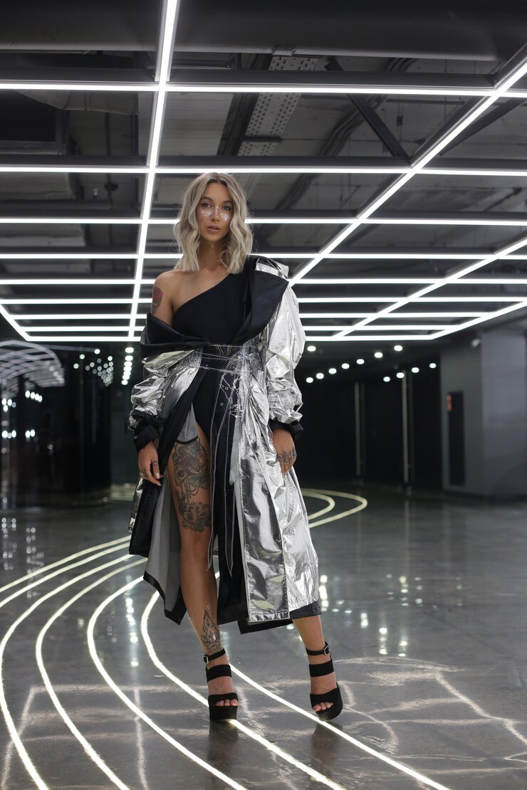 Garota atraente modelo em uma capa de prata brilhante com tatuagens nos pés fica em ambientes fechados com lâmpadas no teto e fita adesiva no chão, tirando fotos de alta qualidade