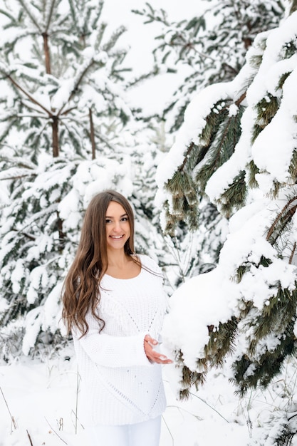 Garota atraente no jumper branco quente na floresta de inverno, olhando para a câmera e sorrindo