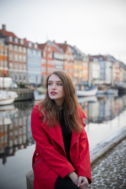 Foto garota atraente sentada, ela é uma turista e está visitando nyhavn em copenhagen, dinamarca. ela está vestindo um casaco vermelho.