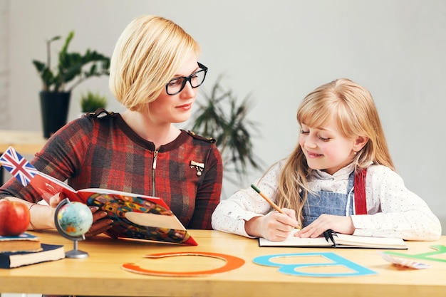 Garota bem-sucedida estudando inglês com mesa de professora loira elegante com processo de aprendizado de livros e letras em sala de aula branca elegante