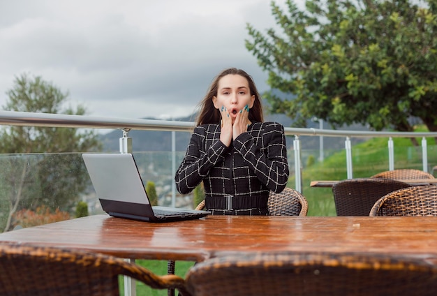 Garota blogueira com laptop está olhando para a câmera segurando as mãos nas bochechas no fundo da vista da natureza