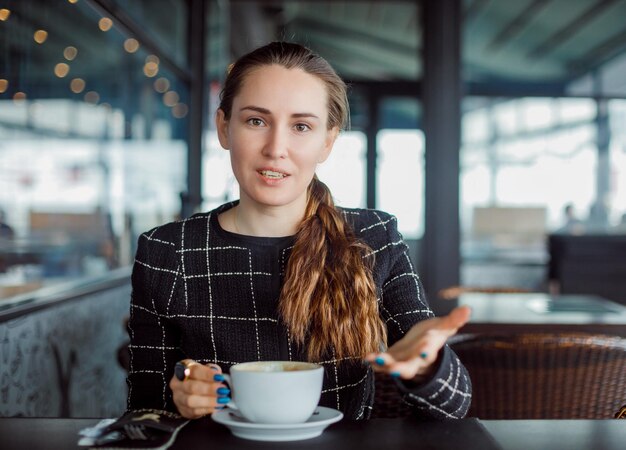 Garota blogueira está olhando para a câmera levantando a mão no café