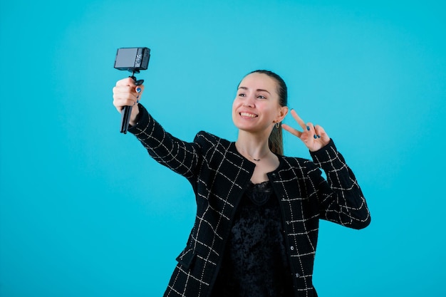 Garota blogueira sorridente está tirando selfie com sua mini câmera em fundo azul