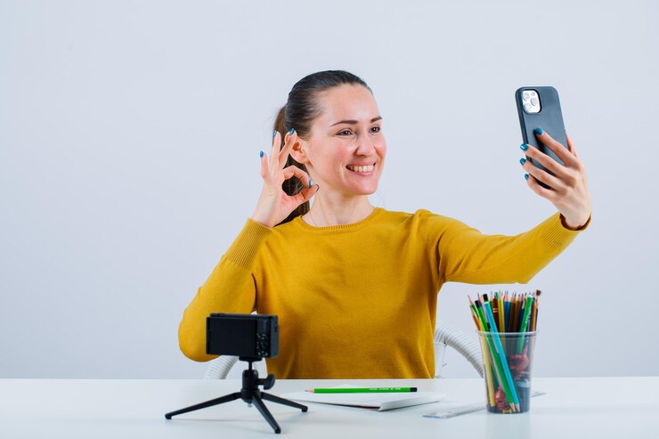 Foto garota blogueira sorridente está tirando selfie com telefone, mostrando um gesto bem no fundo branco