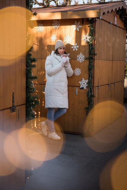 Garota bonita vestida com uma jaqueta branca e um chapéu branco a garota sorri xícara de café na mão casa de verão de madeira garota se alegra emoções