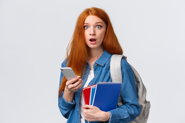 Garota caloura com cabelo vermelho comprido, não sabe onde sua classe, olhando questionada e confusa, olha preocupada, segurando smartphone, mochila e cadernos, sem noção para onde vai, branca