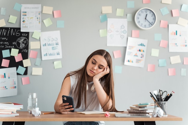 Garota cansada e entediada com um telefone sentado à mesa em um fundo de parede cinza com adesivos. Conceito de aprendizagem.