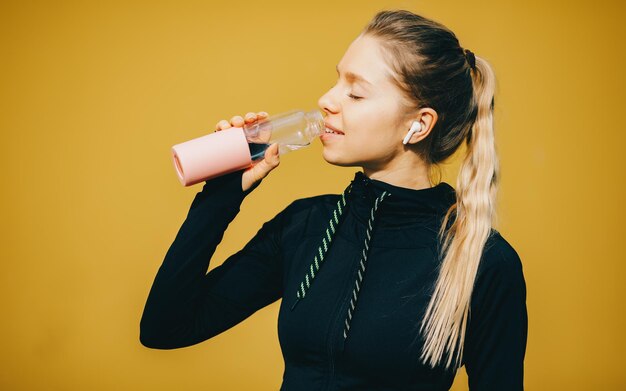 Garota caucasiana atraente em fundo amarelo isolado com fones de ouvido sem fio em traje esportivo faz treino e água potável