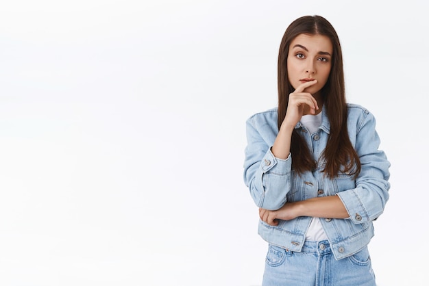 Foto garota caucasiana séria, cética, séria, pensativa, com jaqueta jeans, tocando o lábio e levantando uma sobrancelha, suspeita, suspeita, suspeita de alguém pensando em procurar solução