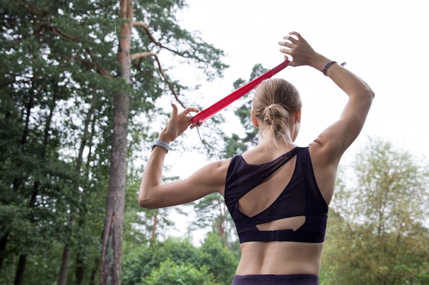 Garota com blusa e leggings treina com corda elástica vermelha em quadras esportivas no parque