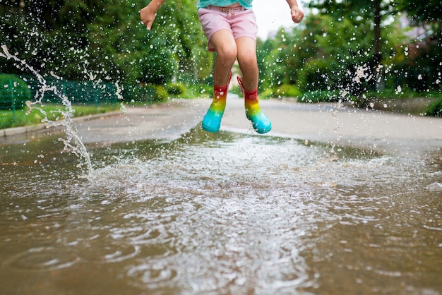 Garota com botas de borracha arco-íris pulando em poças perto de casa