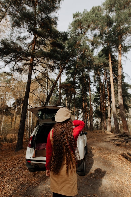 Foto garota com cabelo comprido fica na floresta de outono no contexto de um grande carro