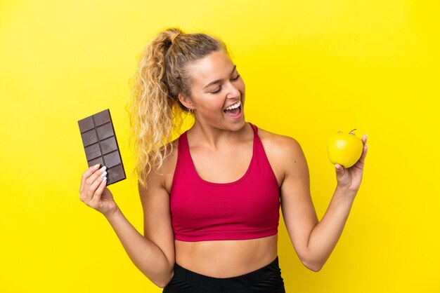 Garota com cabelo encaracolado isolada em fundo amarelo, tomando um tablete de chocolate em uma mão e uma maçã na outra