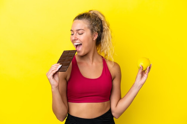 Garota com cabelo encaracolado isolada em fundo amarelo, tomando um tablete de chocolate em uma mão e uma maçã na outra