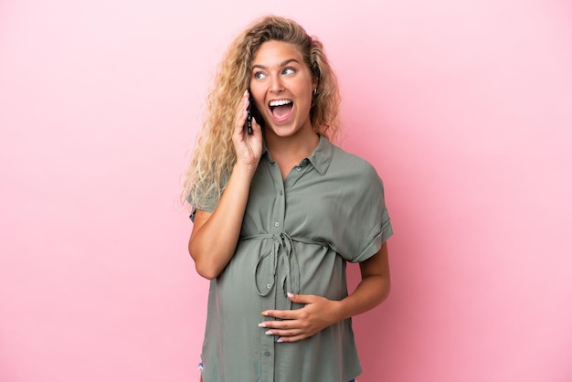 Garota com cabelo encaracolado isolada em fundo rosa grávida e usando telefone celular com expressão de surpresa