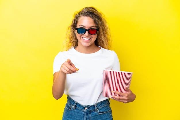 Garota com cabelo encaracolado isolado em fundo amarelo com óculos 3d e segurando um grande balde de pipocas enquanto aponta para a frente