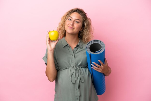 Garota com cabelo encaracolado isolado em um fundo rosa grávida segurando uma maçã e indo para as aulas de ioga