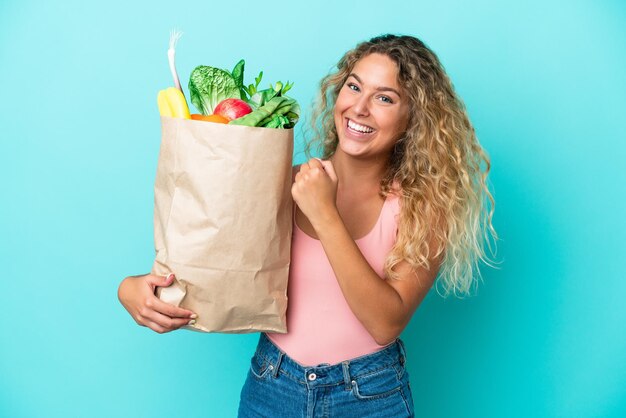 Garota com cabelo encaracolado segurando uma sacola de compras isolada em fundo verde comemorando uma vitória