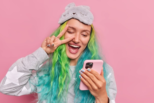 Garota com cabelo tingido faz gesto de paz no olho segura celular leva selfie usa roupa de dormir máscara de dormir na testa posa rosa