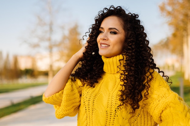 Garota com cachos escuros corrigindo-os com um lindo sorriso olha para longe no parque outono